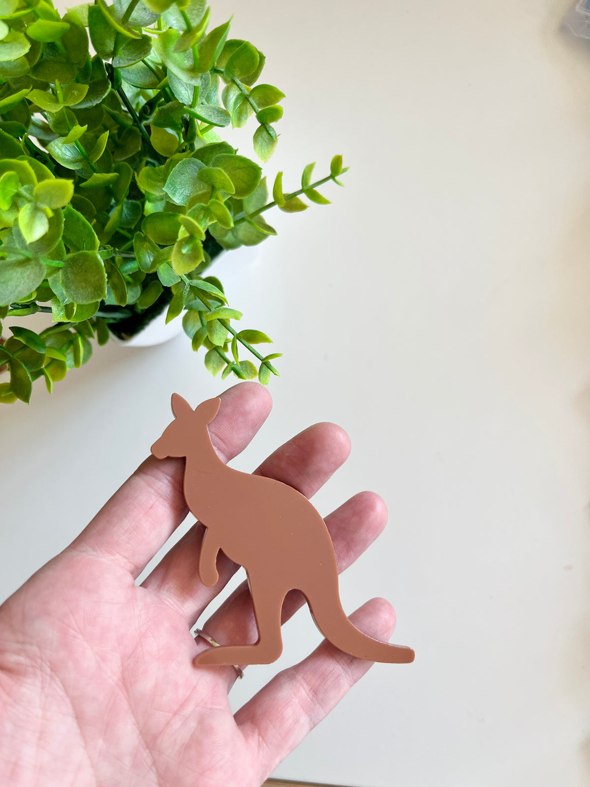 Hand holding a smooth matte brown kangaroo-shaped cutout with green potted leaves on a light background