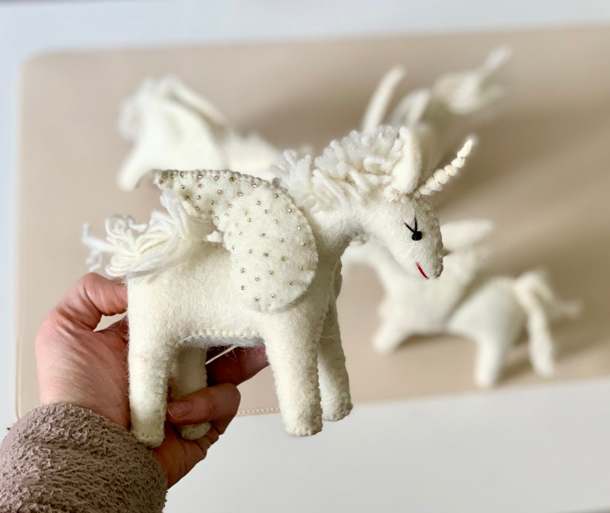 Hand-stitched white felt unicorn toy with a beaded wing and closed eyes held in hand with similar toys in background