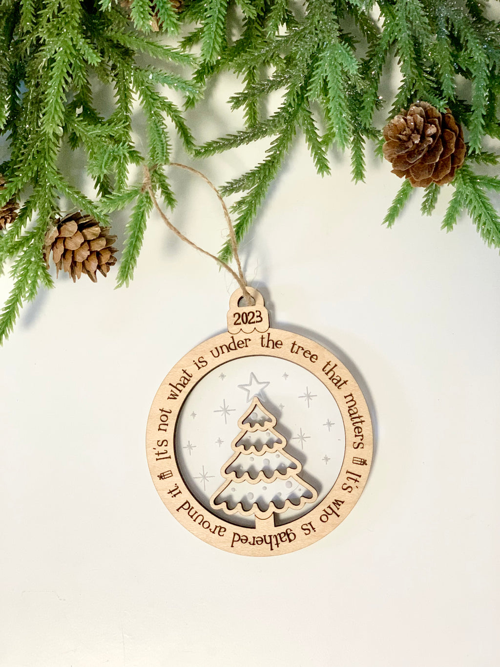 Wood and acrylic Christmas ornament with engraved tree and 2023 date hanging from green pine branch with pinecones