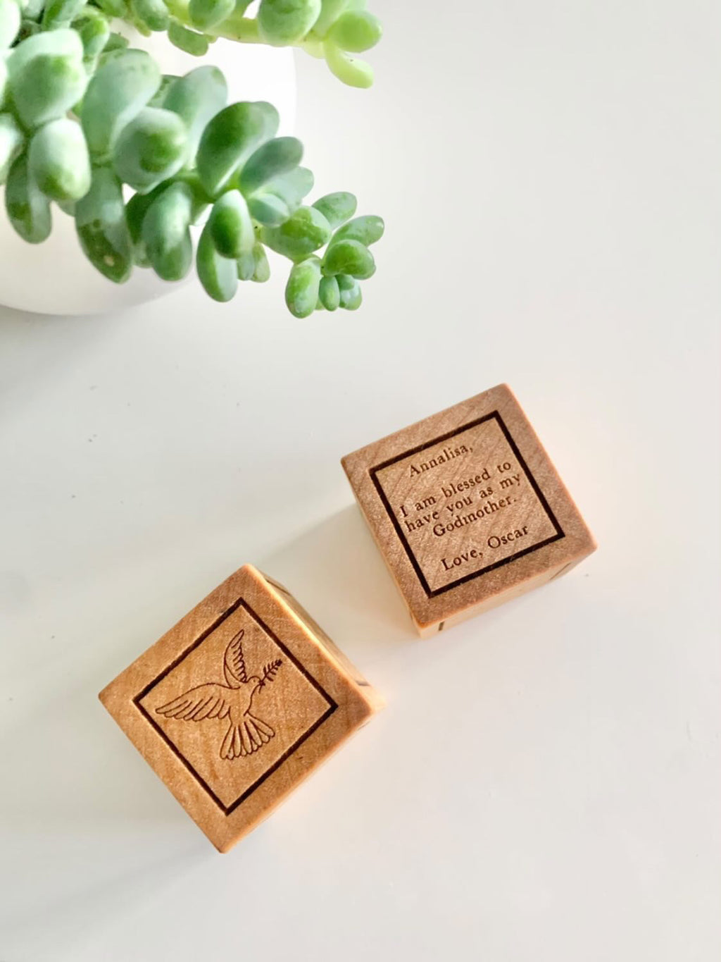 Two personalized wooden baptism blocks with engraved dove and custom message on white surface next to green succulent