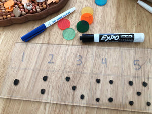 Clear plastic board with numbers one to five, black counters below each number, dry erase markers, and colorful discs on wood surface