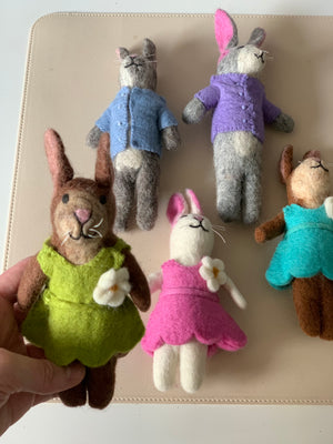 Five handmade felt bunny dolls dressed in colorful outfits on a beige surface with one held in hand
