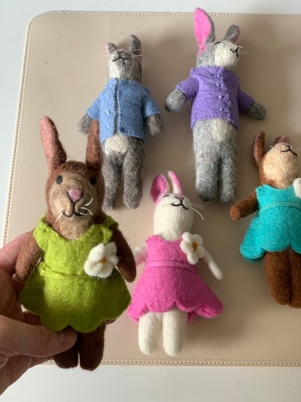 Five handmade felt bunny dolls dressed in colorful outfits on a beige surface with one held in hand