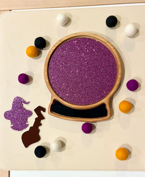 Wooden sensory tray shaped like a snow globe with purple glitter interior and felt balls in purple, black, white, and orange surrounding it