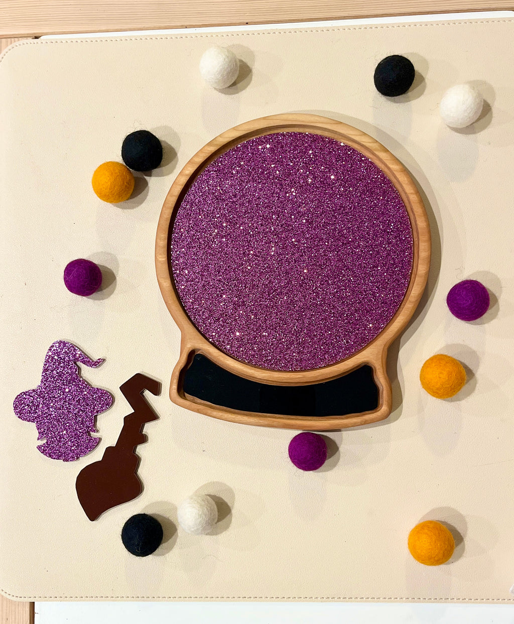 Wooden sensory tray shaped like a snow globe with purple glitter interior and felt balls in purple, black, white, and orange surrounding it