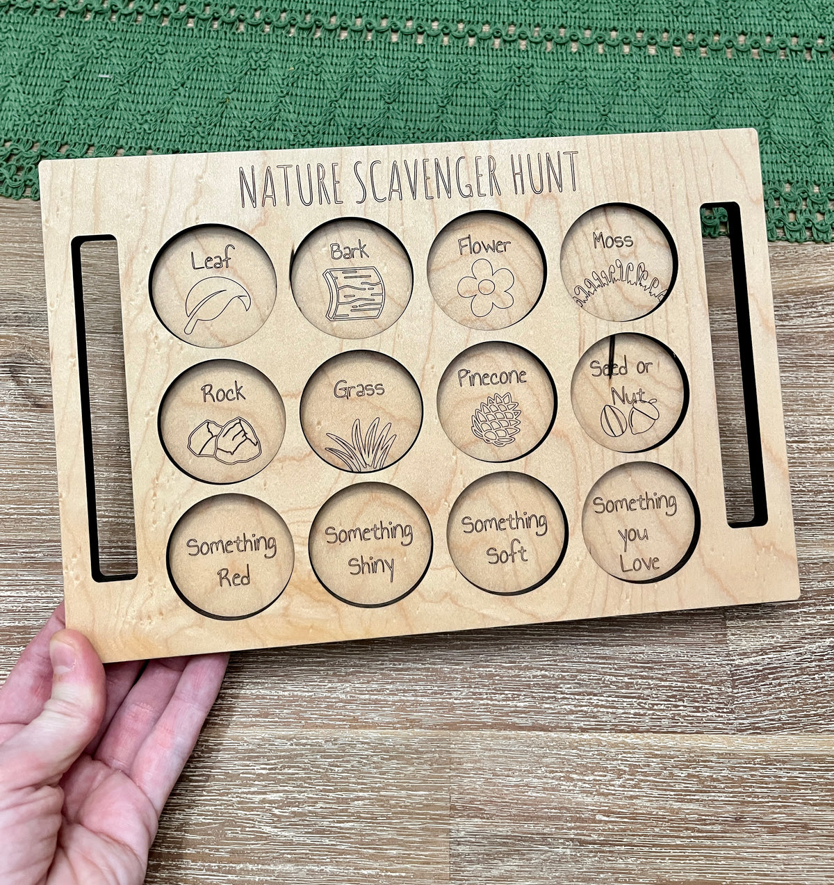 Natural wooden Nature Scavenger Hunt board with engraved circles and nature-themed words and illustrations held by hand on wood surface