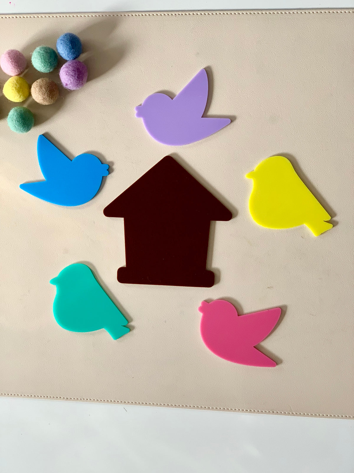 Colorful bird-shaped cutouts surrounding a brown house-shaped cutout with pastel pom-poms nearby on a beige surface