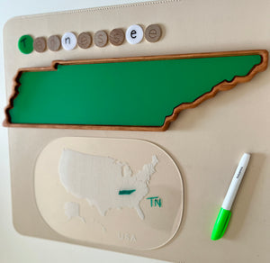 Green wooden tray shaped like Tennessee with letter tokens spelling Tennessee and a dry erase marker on beige surface