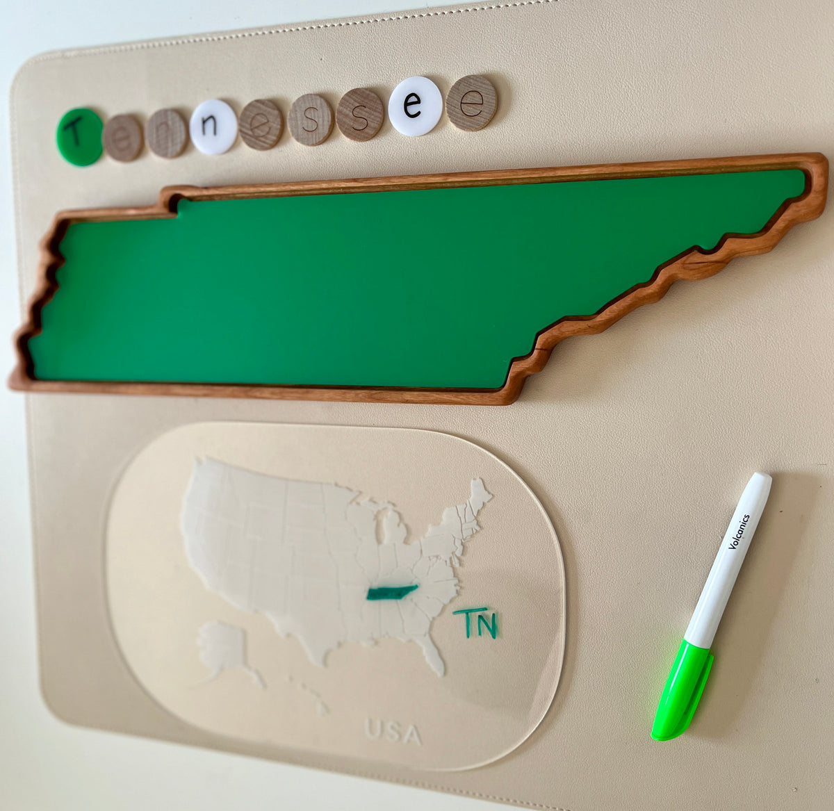 Green wooden tray shaped like Tennessee with letter tokens spelling Tennessee and a dry erase marker on beige surface