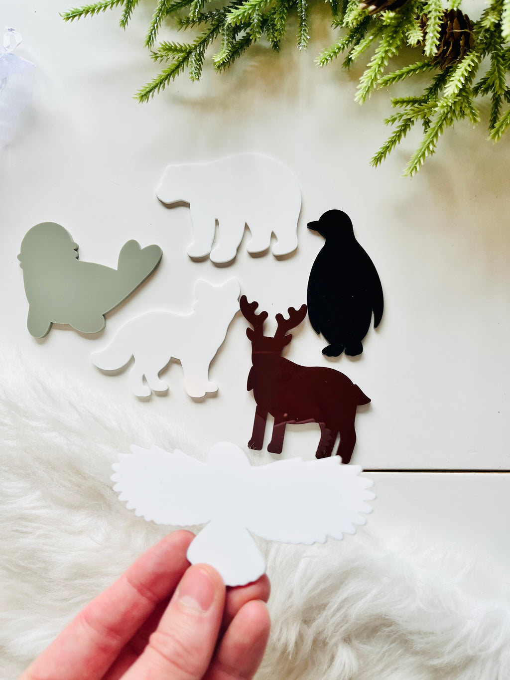 Hand holding white bird-shaped acrylic cutout near flat acrylic animal shapes including bear, penguin, moose, and seal on white surface