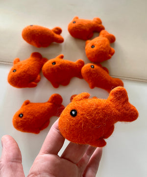 Hand holding small bright orange felt fish toy with black eyes above several similar fish on light surface