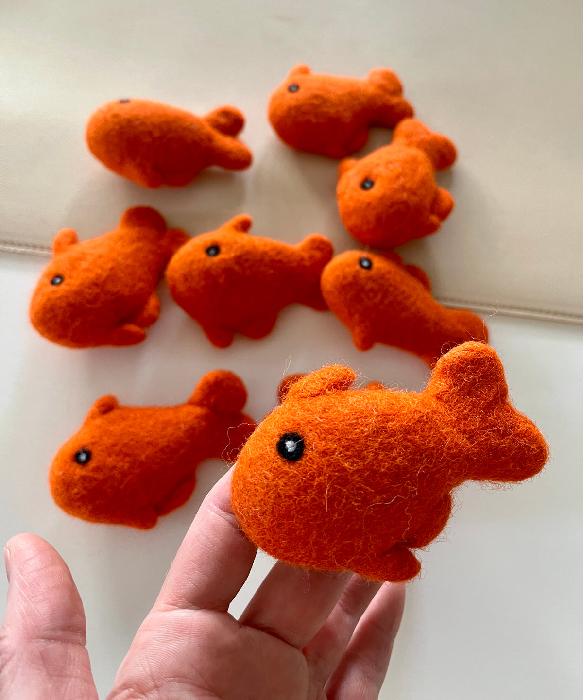 Hand holding small bright orange felt fish toy with black eyes above several similar fish on light surface