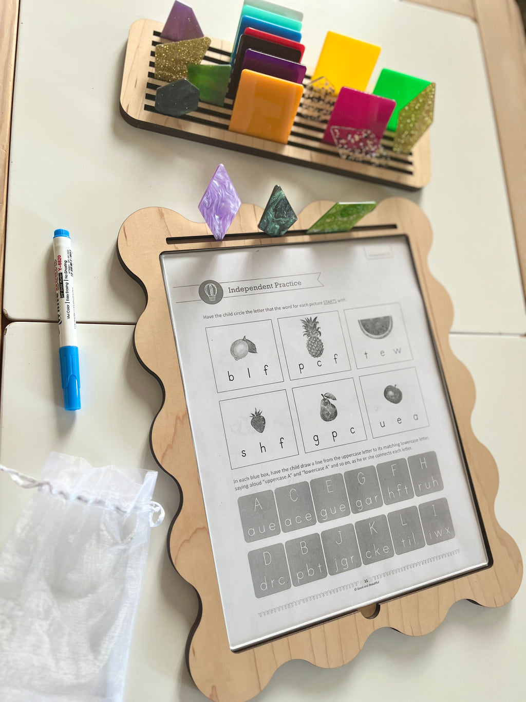 Wooden-framed activity board with colorful translucent shapes clipped on top and a dry erase marker beside a white pouch