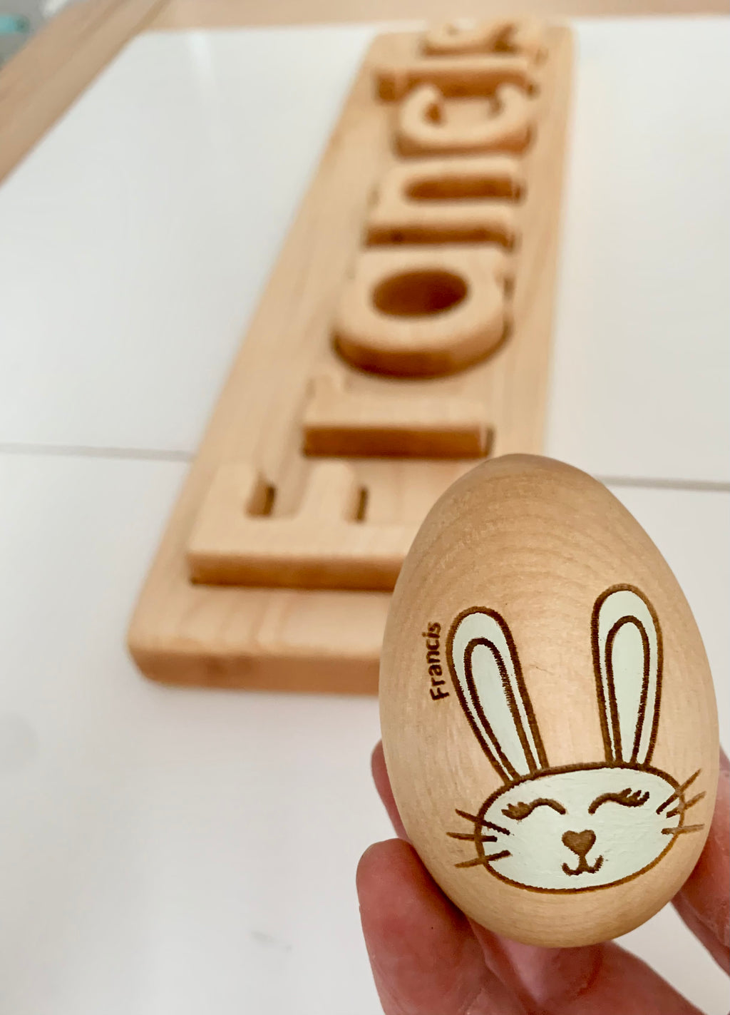 Natural wooden personalized Easter egg with bunny face illustration and name engraved held over wooden name sign on white surface