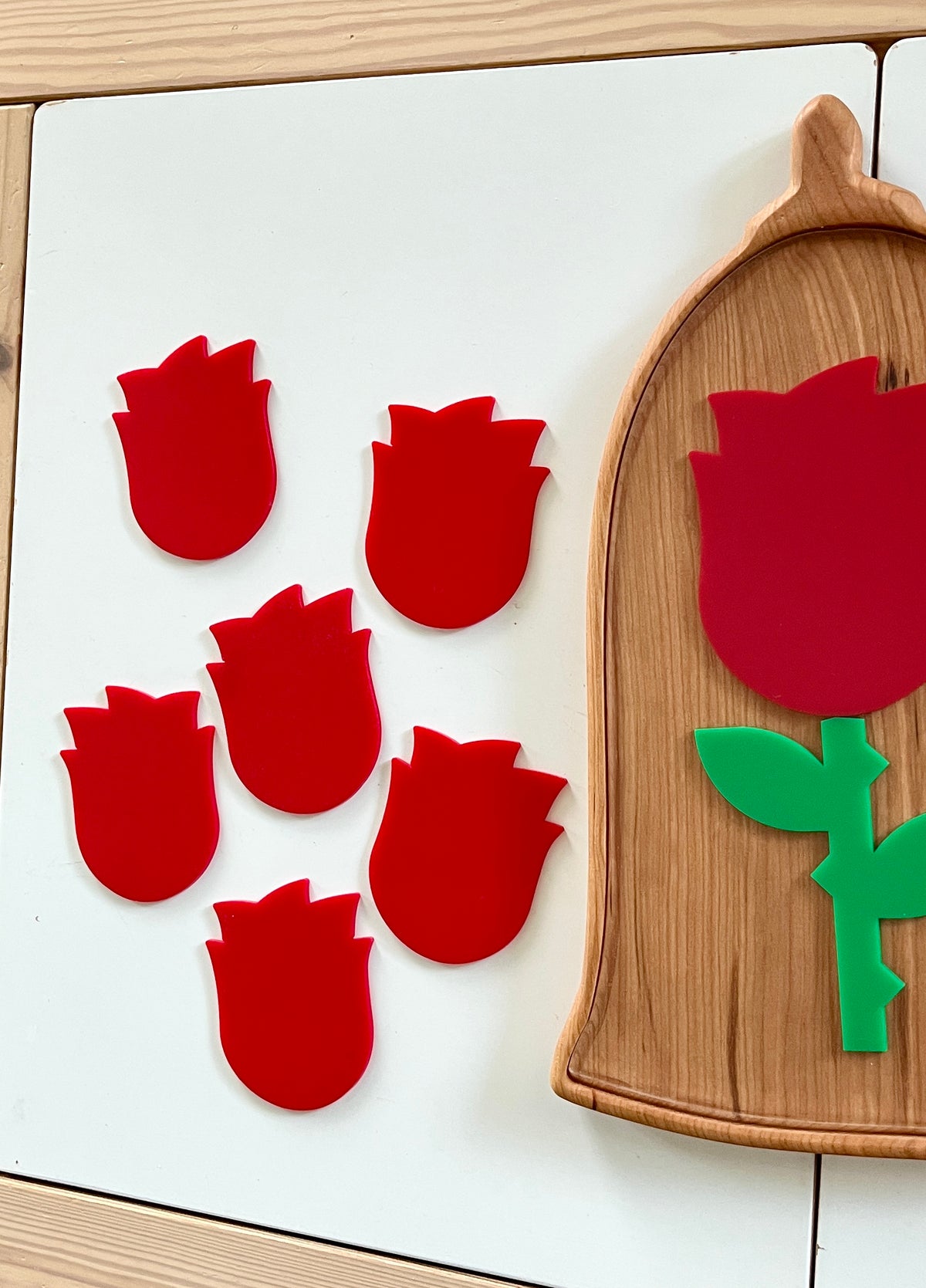Six small red tulip-shaped acrylic pieces next to a wooden tray holding a larger red tulip with green stem and leaves