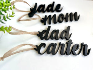 Four black acrylic name tags reading jade, mom, dad, and carter with tan hanging ribbons on white surface