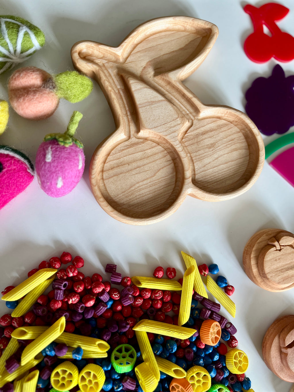 Wooden cherry-shaped tray with colorful pasta pieces, felt fruit decorations, and wooden apple trays on white surface