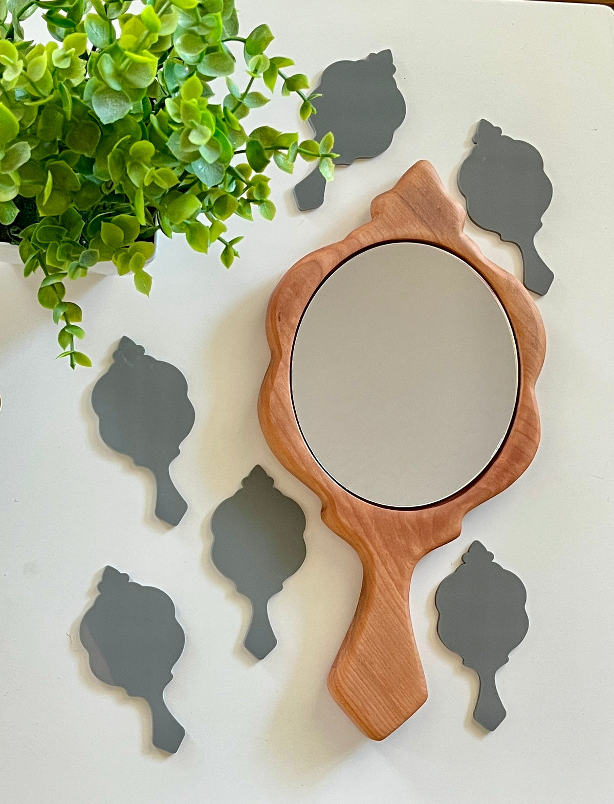 Wooden framed handheld mirror surrounded by six small gray silhouette cutouts on white surface next to green plant
