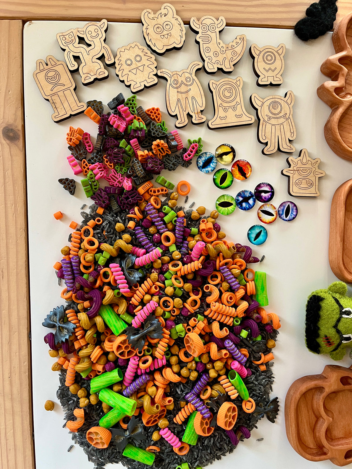 Wooden monster tokens with colorful spiral pasta, plastic monster eye pieces, and wooden trays on light surface