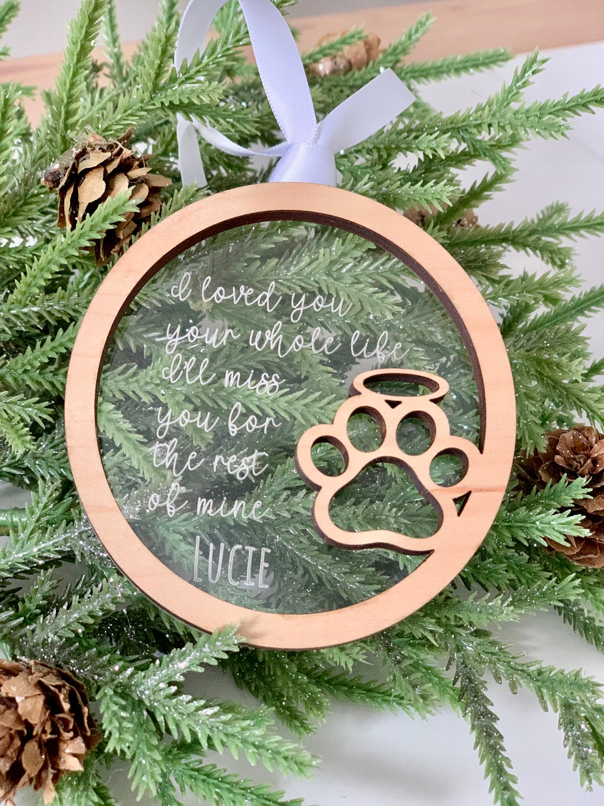 Round wooden ornament with engraved paw print and heartfelt message against pine branches with pine cones