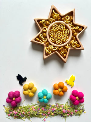 Star-shaped wooden tray with yellow pasta wheels surrounded by felt flowers, bees, and scattered green herbs on white surface