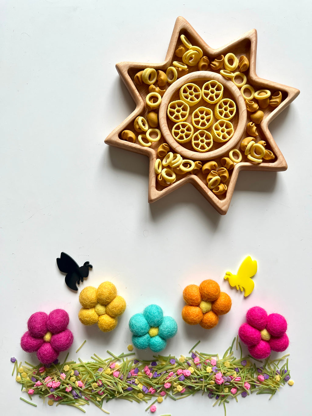 Star-shaped wooden tray with yellow pasta wheels surrounded by felt flowers, bees, and scattered green herbs on white surface