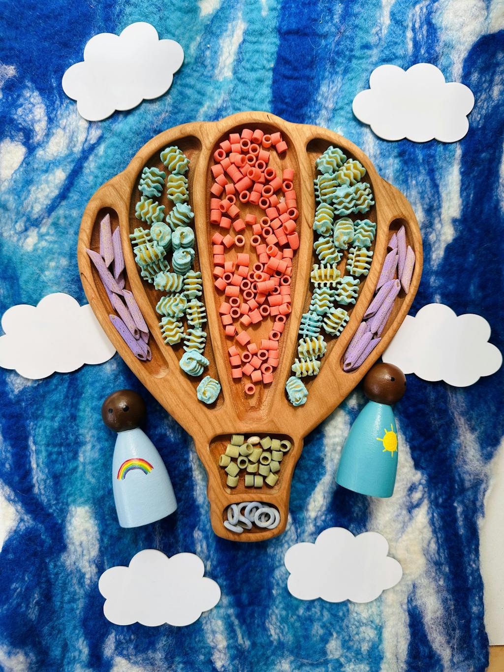 Wooden hot air balloon sensory tray filled with colorful pasta shapes on blue and white felt with wooden peg dolls and paper clouds