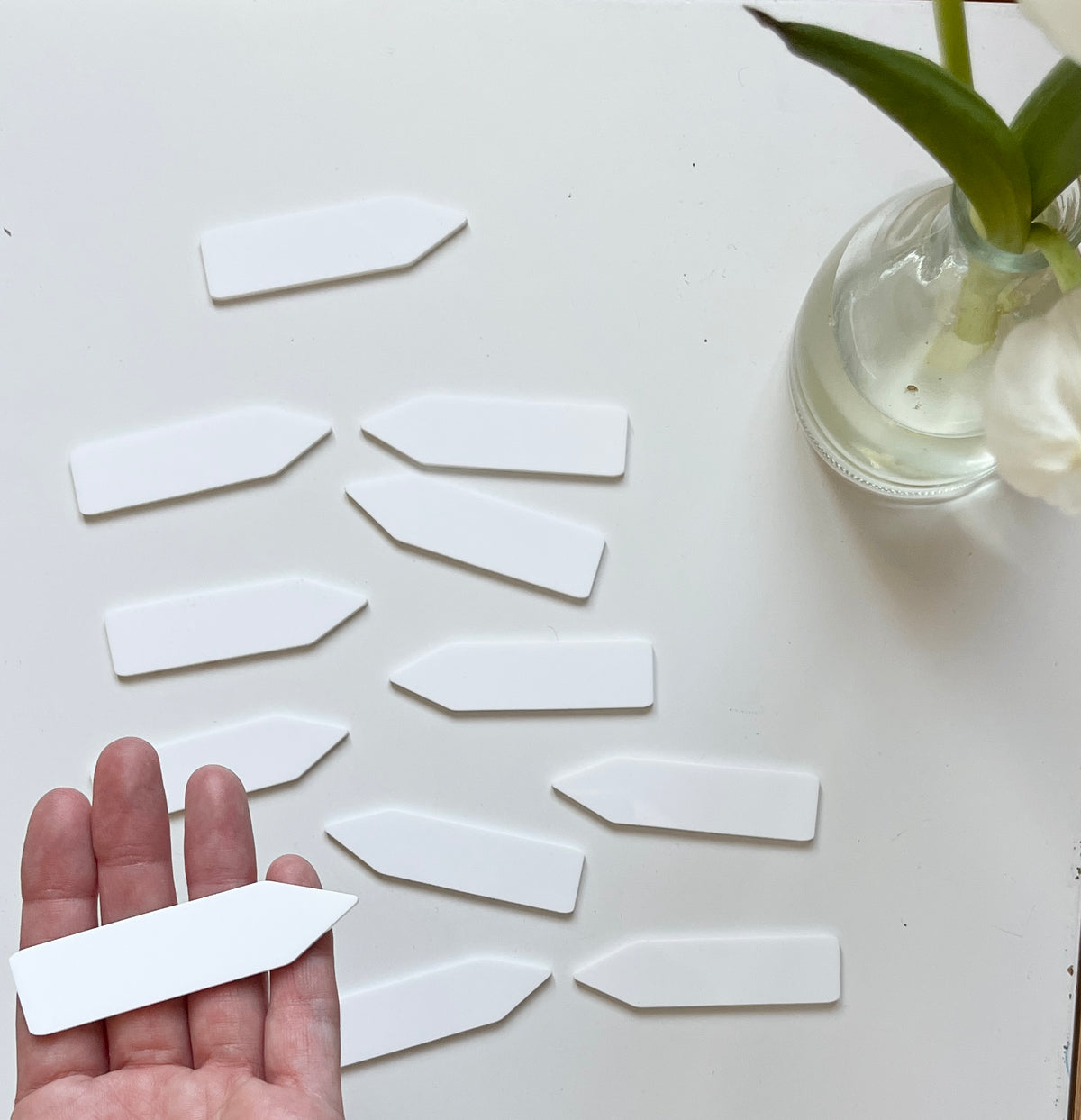 Hand holding a white arrow-shaped plastic plant label with several more scattered on a white surface near a glass vase with green plant leaves