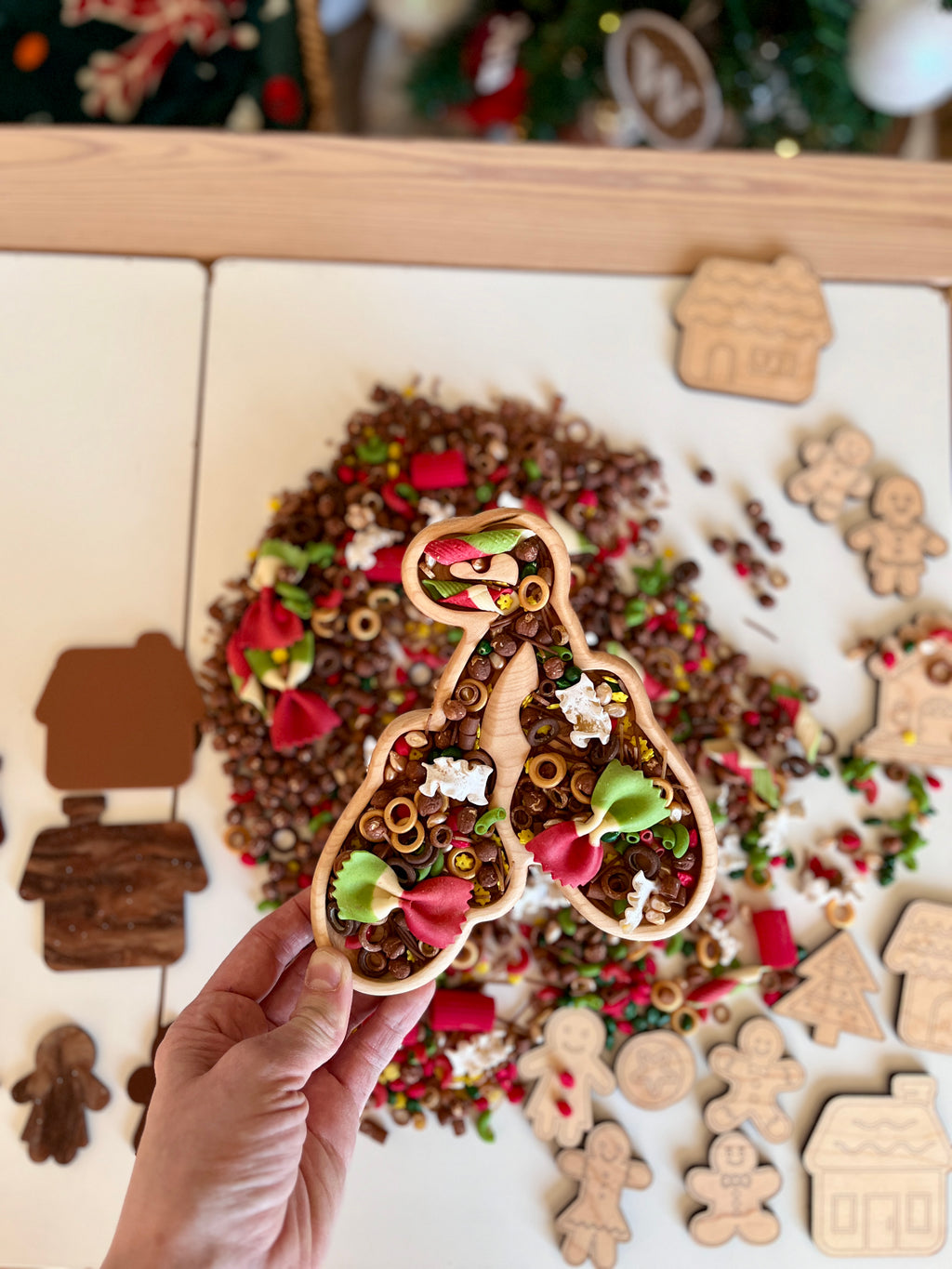 Hand holding wooden mitten-shaped tray filled with pasta shapes and colorful beads on a white table with wooden holiday cutouts nearby