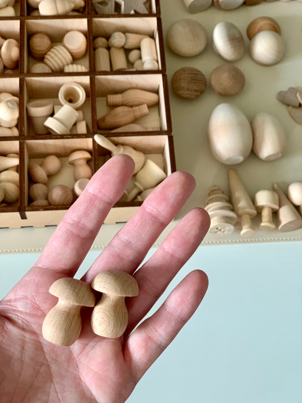 Hand holding two small smooth wooden mushroom shapes with assorted unfinished wooden toys and shapes in background