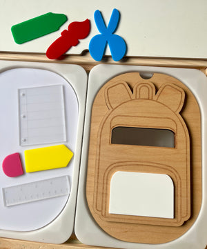Wooden backpack-shaped puzzle piece with cutouts beside colorful plastic stationery shapes and clear page and ruler pieces on white trays