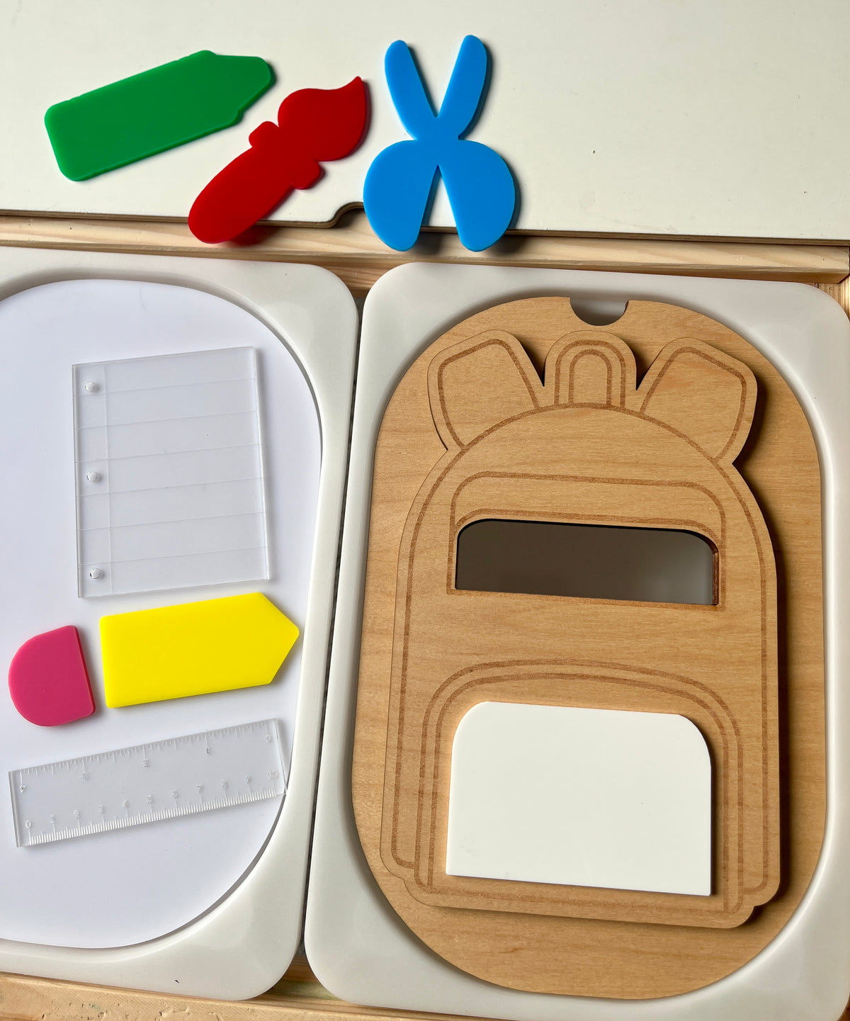 Wooden backpack-shaped puzzle piece with cutouts beside colorful plastic stationery shapes and clear page and ruler pieces on white trays