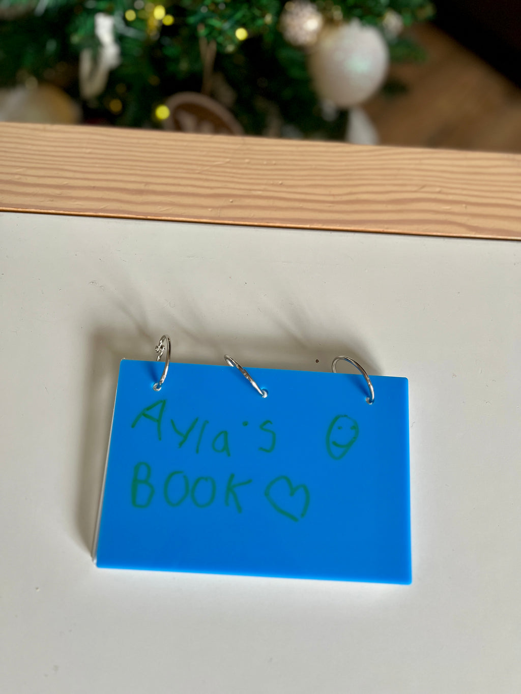 Small blue plastic book with handwritten green text and three metal rings on white surface near Christmas tree