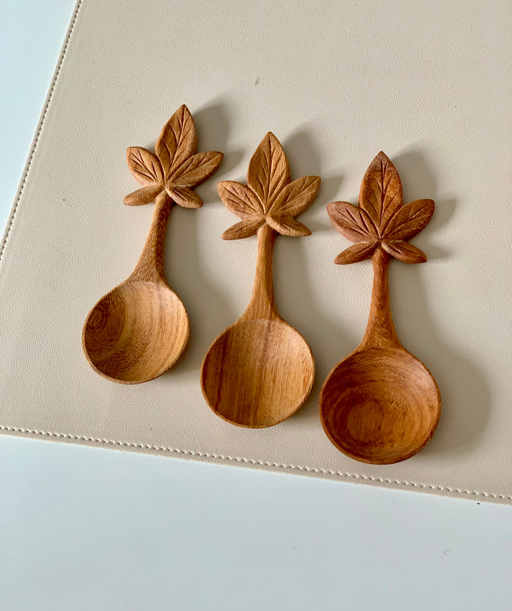 Three carved wooden spoons with handles shaped like maple leaves resting on a beige textured surface