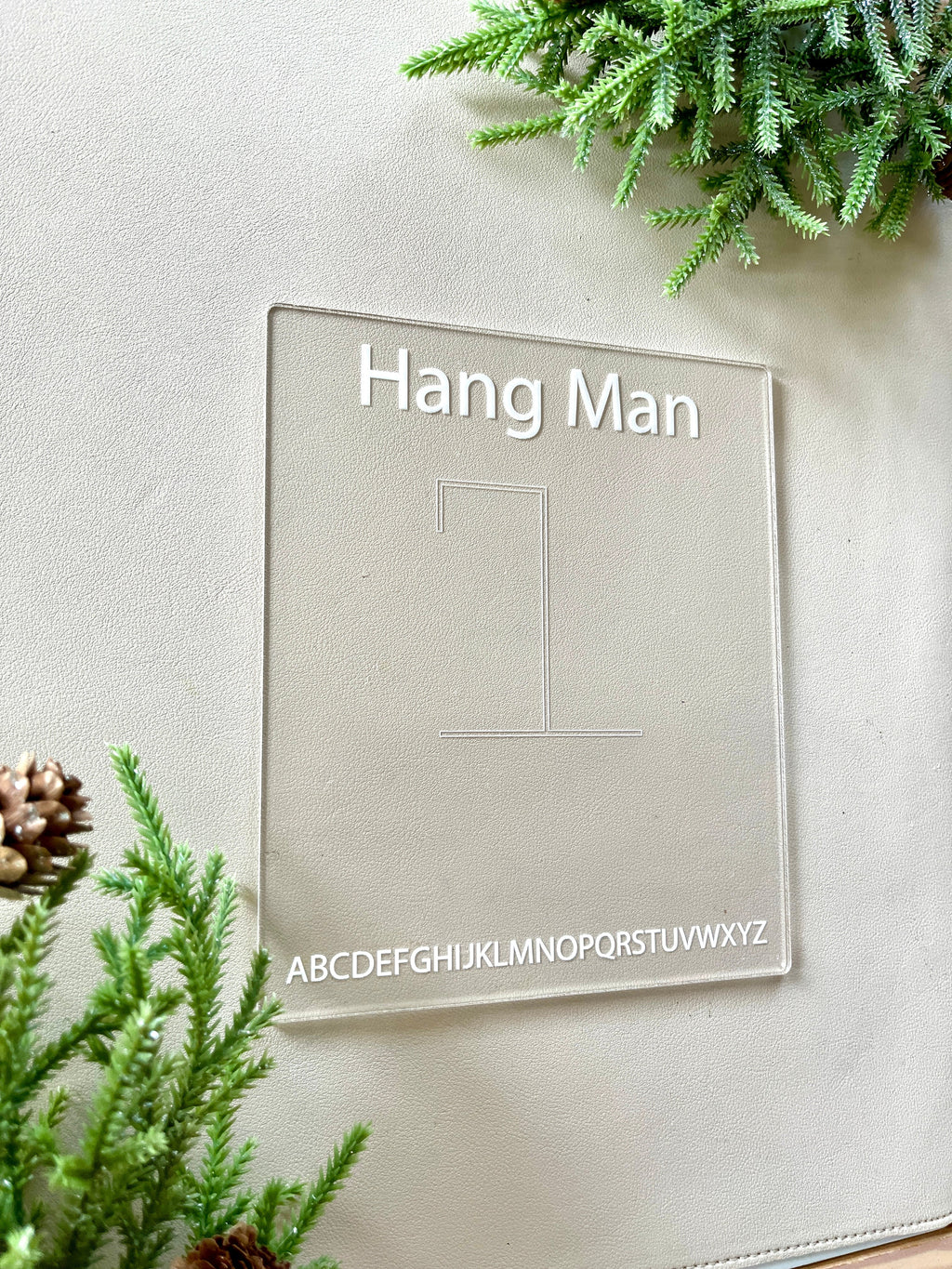 Clear acrylic board engraved with hangman game layout and alphabet letters, surrounded by green foliage and pinecones