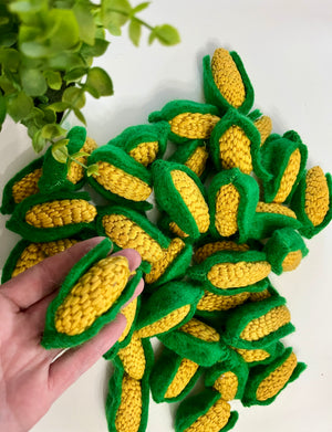 Hand holding knitted yellow corn cob with green husk, pile of similar knitted corn cobs on white surface near green plant