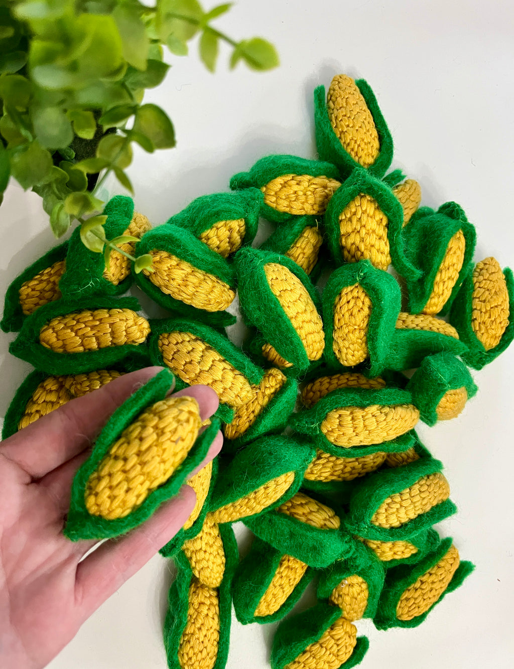 Hand holding knitted yellow corn cob with green husk, pile of similar knitted corn cobs on white surface near green plant