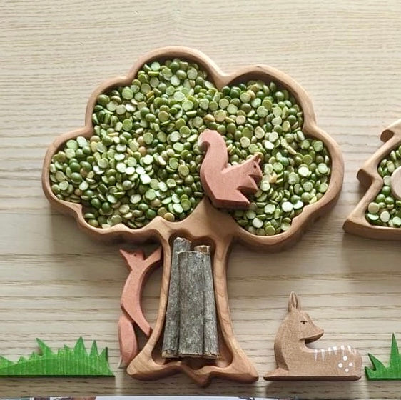Tree-shaped wooden sensory tray filled with green split peas and small wooden animal figures on light wood surface