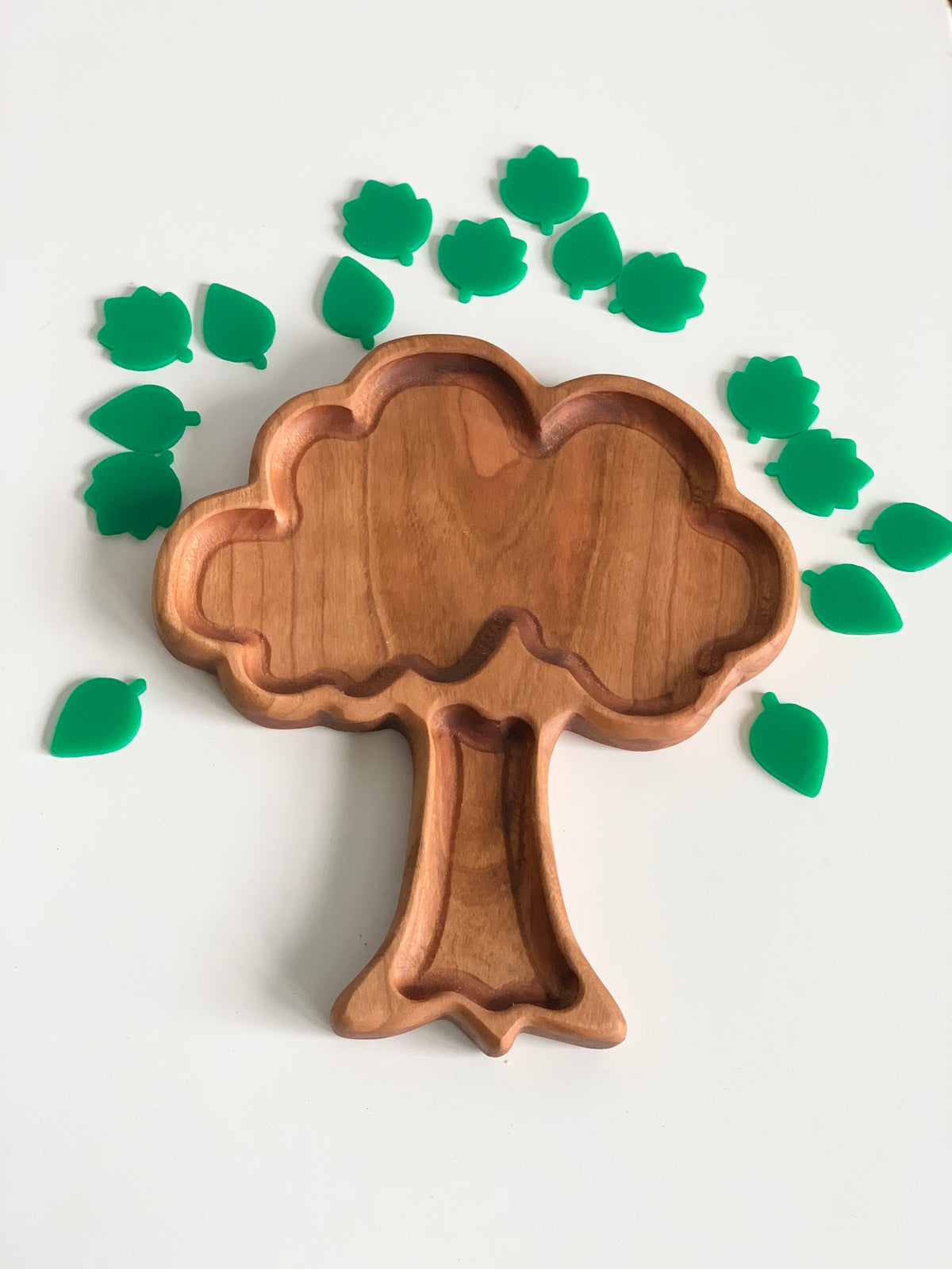 Wooden tree-shaped tray with multiple small green leaf-shaped tokens arranged around it on a white surface