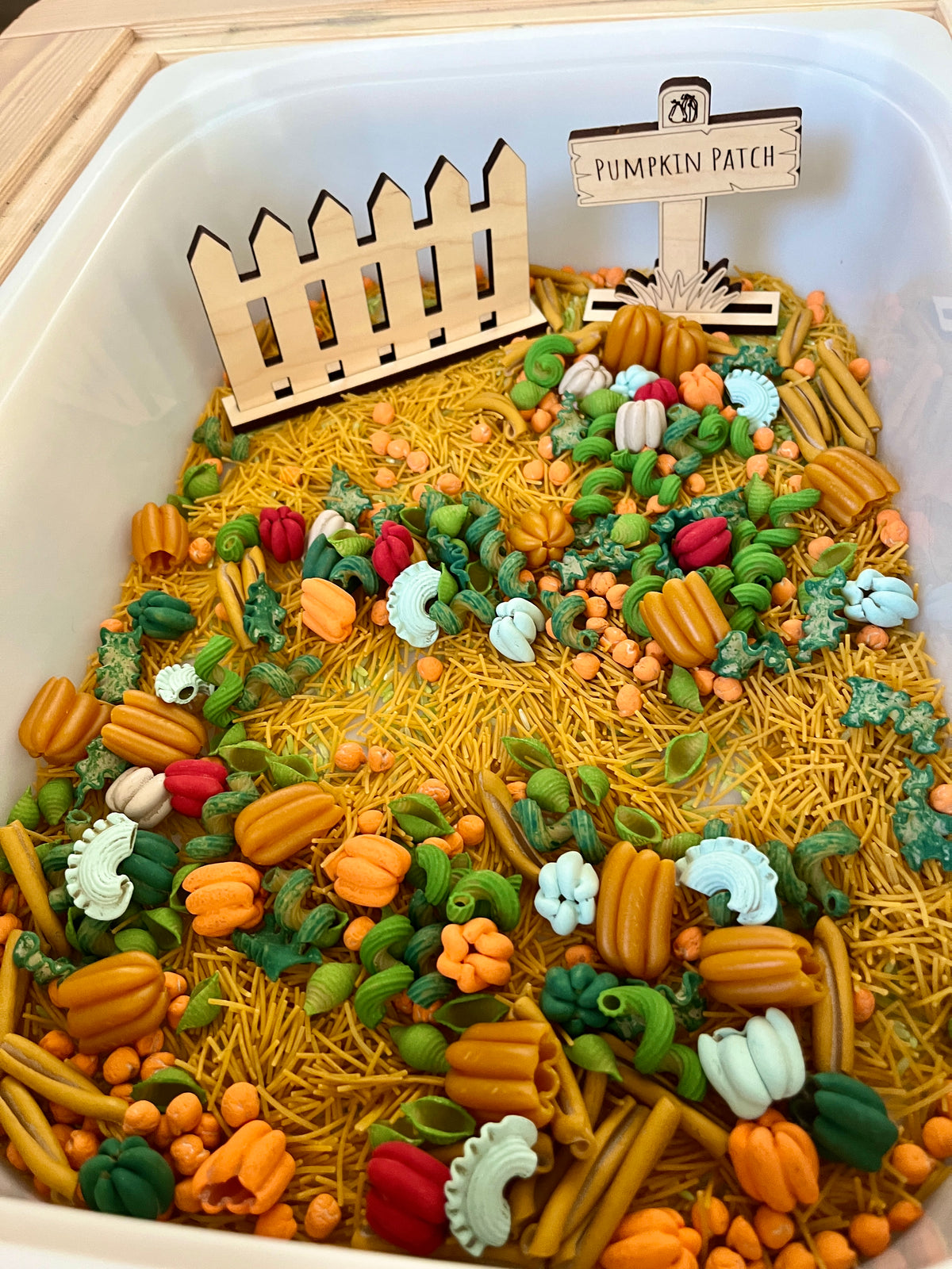 White container filled with colorful pasta shapes and dry noodles, decorated with wooden fence and pumpkin patch sign
