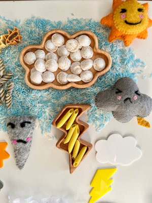 Wooden cloud and lightning trays filled with shells and yellow pasta pieces on blue and white rice with felt weather shapes