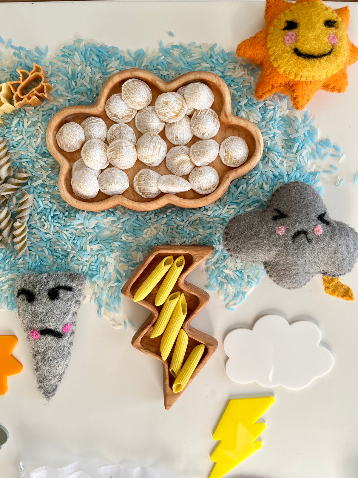 Wooden cloud and lightning trays filled with shells and yellow pasta pieces on blue and white rice with felt weather shapes