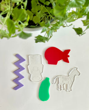 Purple zigzag, clear robot figure, red fish, green bottle, and clear zebra acrylic shapes on white surface with green leaves above