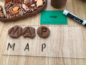 Clear board with uppercase letter tiles spelling MAP alongside matching word card and sensory play materials on wooden surface