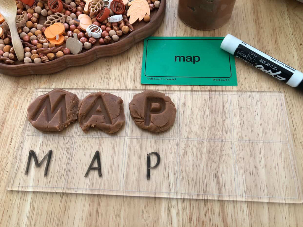 Clear board with uppercase letter tiles spelling MAP alongside matching word card and sensory play materials on wooden surface