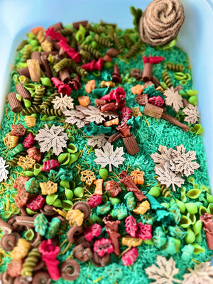 Blue container filled with green rice, colorful pasta shapes, wooden leaves, and a felt pinecone decoration