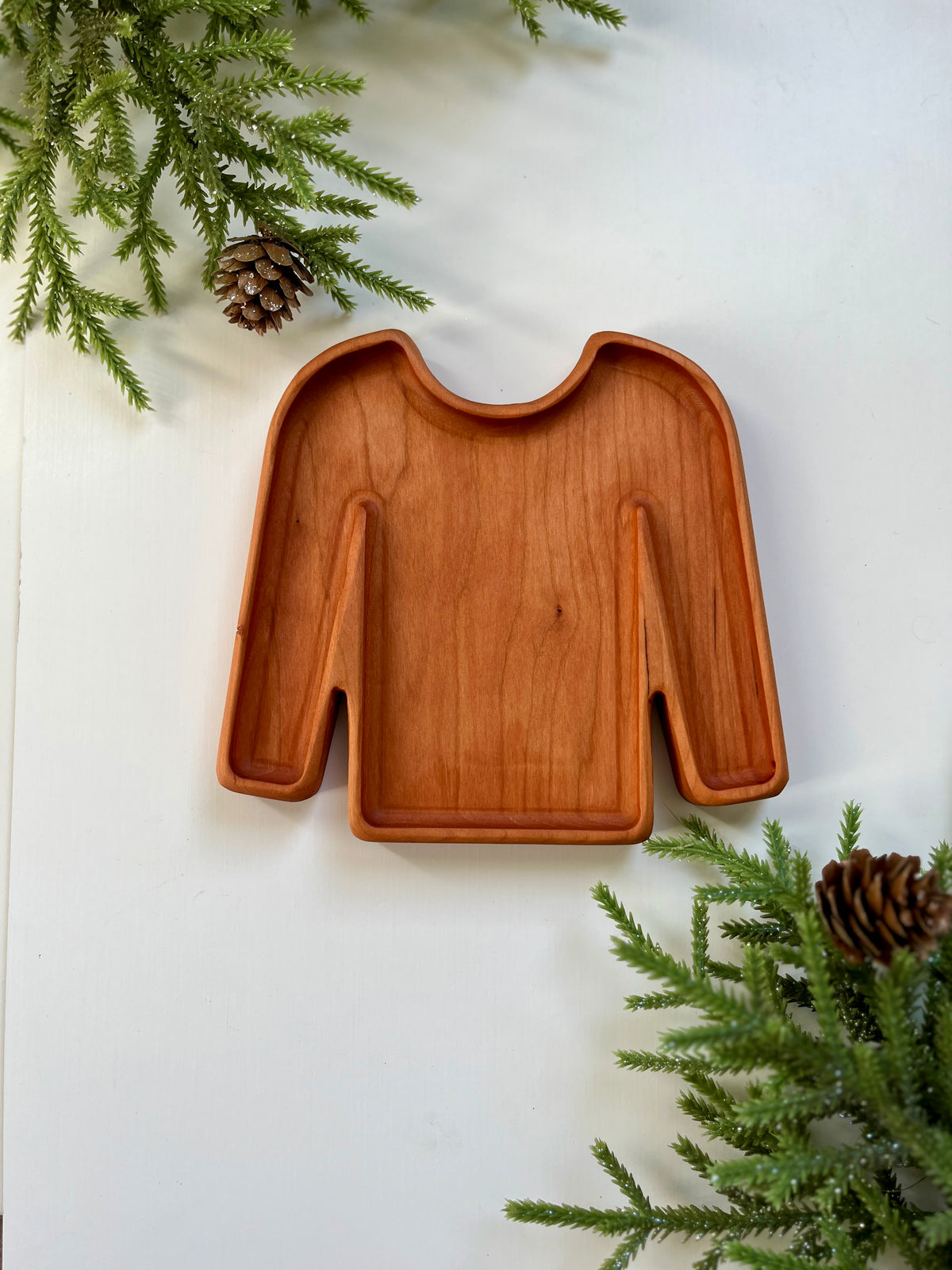 Wooden tray carved in the shape of a long-sleeve sweater surrounded by pine branches and pine cones on white surface