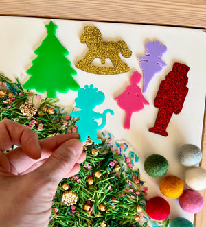 Hand holding blue dinosaur cutout above green, red, purple, pink, and gold holiday-themed acrylic shapes with mixed sensory materials
