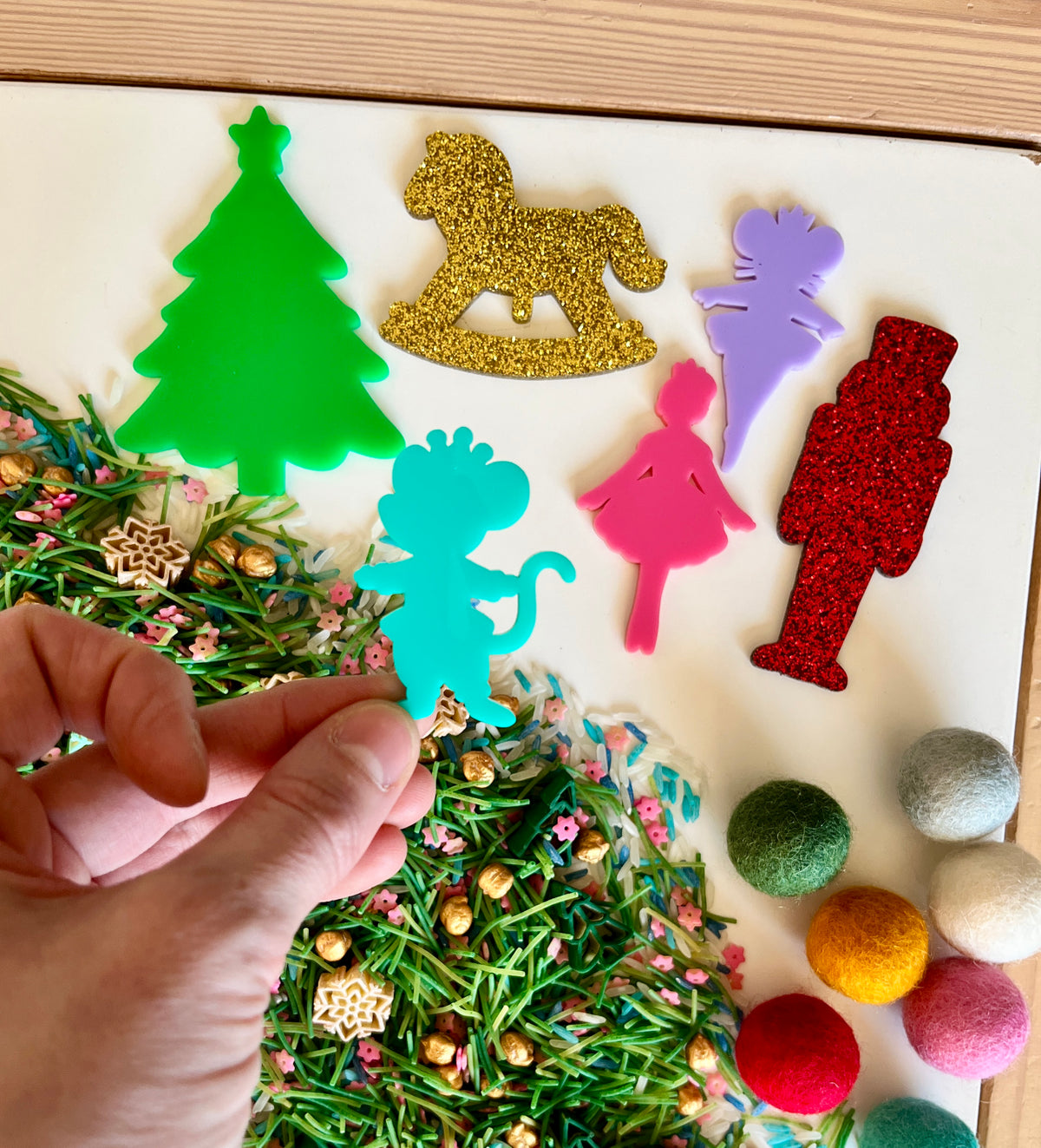 Hand holding blue dinosaur cutout above green, red, purple, pink, and gold holiday-themed acrylic shapes with mixed sensory materials