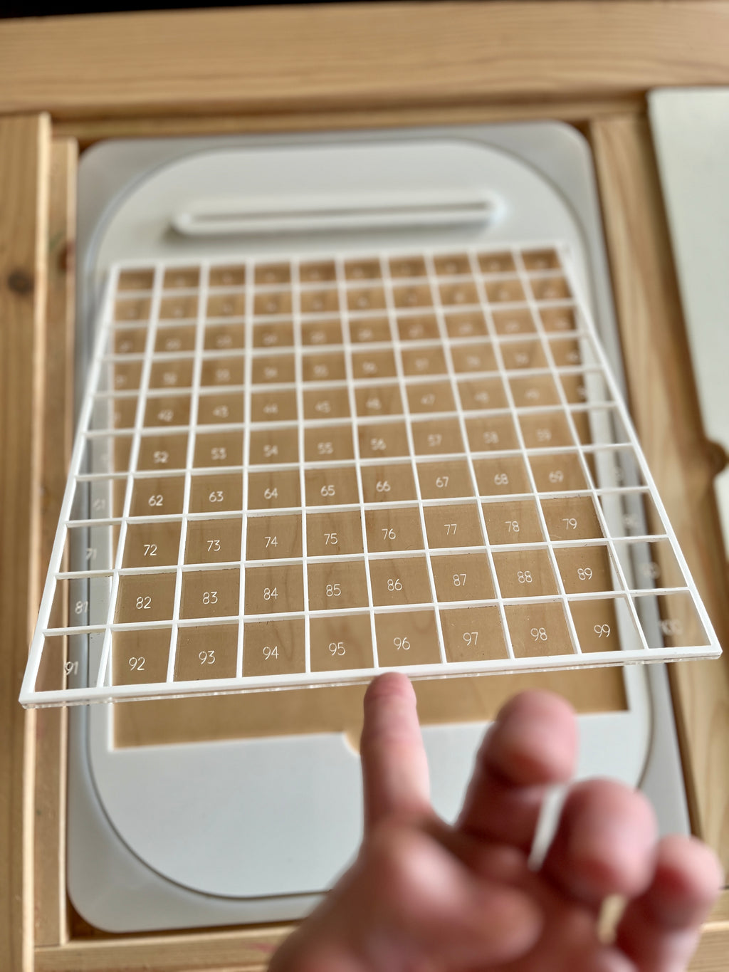 Hand holding a white plastic grid above a numbered 100 square chart on a beige wooden frame background